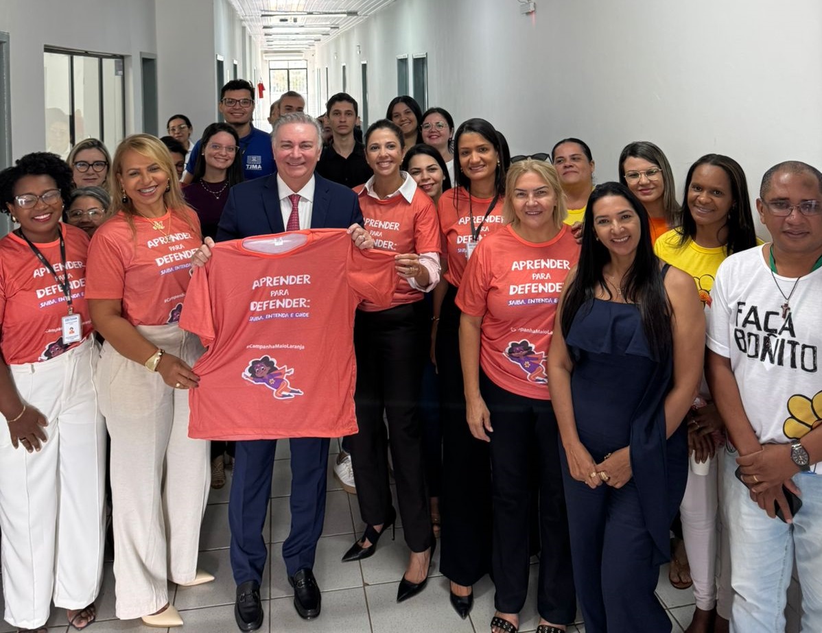 A imagem mostra um grupo de aproximadamente 20 pessoas reunidas em um corredor de um prédio com paredes brancas e piso claro. A iluminação parece vir do teto, com luminárias embutidas visíveis.

No centro da imagem, um homem de terno escuro e gravata vermelha segura uma camiseta laranja desdobrada, exibindo a frase "APRENDER PARA DEFENDER:" em letras brancas e uma ilustração de uma criança. Duas mulheres ao lado dele, uma à sua esquerda e outra à sua direita, também seguram a camiseta. A mulher à esquerda veste uma camiseta laranja semelhante com a mesma frase e calças brancas, enquanto a mulher à direita veste uma camiseta laranja com a mesma frase e calças pretas.

Várias outras pessoas ao redor vestem camisetas laranjas com a mesma estampa. Algumas pessoas na parte de trás vestem outras cores, como preto, branco e amarelo. Há uma variedade de tons de pele e estilos de cabelo entre as pessoas no grupo.

A atmosfera geral parece positiva e de apoio, sugerindo um evento ou iniciativa de grupo. As camisetas idênticas indicam que eles podem estar participando de uma campanha ou evento específico relacionado ao tema "APRENDER PARA DEFENDER:".

Ao fundo, à esquerda, há uma porta azul e, no final do corredor, vê-se uma área mais iluminada que parece ser a entrada ou saída do prédio, com luz natural entrando. Há também outras portas ao longo do corredor.