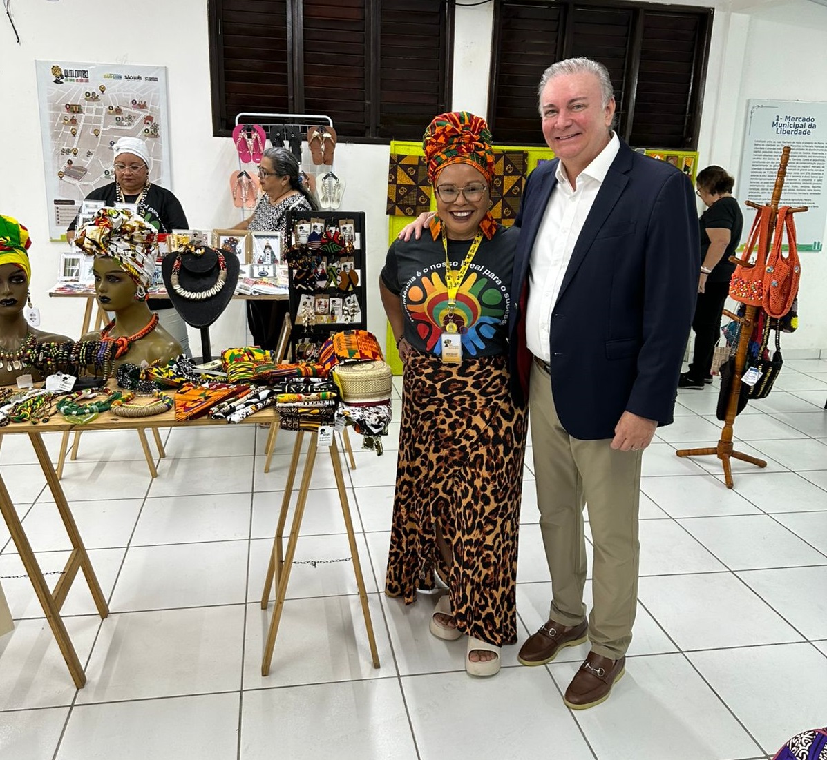 A imagem mostra duas pessoas em pé, posando para uma foto em um ambiente interno que parece ser uma feira de artesanato ou uma loja.

À esquerda, há uma mulher com pele morena, usando óculos, um turbante vibrante em tons de laranja e verde, e uma camiseta preta com uma estampa colorida que inclui figuras e texto em português. Ela veste uma saia longa com estampa de leopardo e sandálias plataforma claras. Ela está sorrindo e tem o braço esquerdo ao redor do homem à sua direita.

À direita, o desembargador Froz Sobrinho:um homem de pele clara, com cabelos grisalhos e um sorriso. Ele veste um blazer azul marinho sobre uma camisa branca e calças cáqui, além de sapatos marrons.

Ao redor deles, há mesas de madeira com diversos itens de artesanato à venda, incluindo colares, pulseiras, tecidos estampados e turbantes em manequins. Ao fundo, outras pessoas podem ser vistas olhando os produtos. Há também pôsteres ou informativos pendurados na parede, um deles parcialmente visível com o título "3 - Mercado Municipal da Liberdade". O chão é claro e parece ser de cerâmica. A iluminação é artificial, vinda do teto.