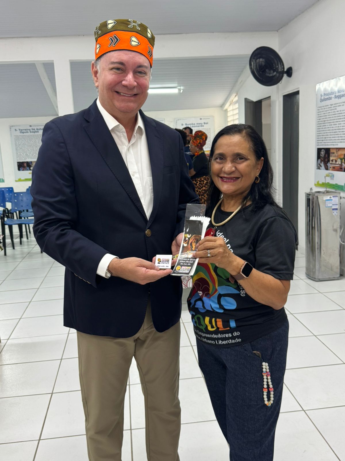 A imagem mostra duas pessoas posando para uma foto em um ambiente interno, que parece ser um espaço de exposição ou evento cultural.

À esquerda, um homem de pele clara com um sorriso amigável veste um blazer azul escuro sobre uma camisa branca e calças cáqui. Ele também usa um acessório de cabeça laranja e marrom com padrões tribais. Ele está segurando um pequeno cartão branco e um folheto colorido em suas mãos, oferecendo-os à mulher ao seu lado.

À direita, uma mulher com pele morena e cabelos escuros está sorrindo para a câmera. Ela veste uma camiseta preta com uma estampa colorida que inclui figuras e texto, e calças jeans escuras. Ela usa um colar de pérolas brancas e um relógio preto no pulso esquerdo. Ela parece estar segurando o folheto que o homem lhe oferece.

Ao fundo, parcialmente visíveis, há pôsteres ou painéis informativos pendurados na parede com texto em português e algumas imagens. Também se veem cadeiras azuis e outras pessoas ao longe. O chão é de cor clara e parece ser de azulejos. A iluminação é artificial, vinda do teto. O ambiente sugere uma atmosfera de celebração cultural ou de apoio ao empreendedorismo local.