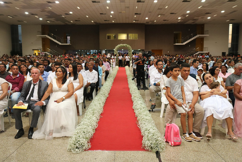 noivos e noivas sentados em cadeiras plásticas em salão dividido por um tapete vermelho ao centro