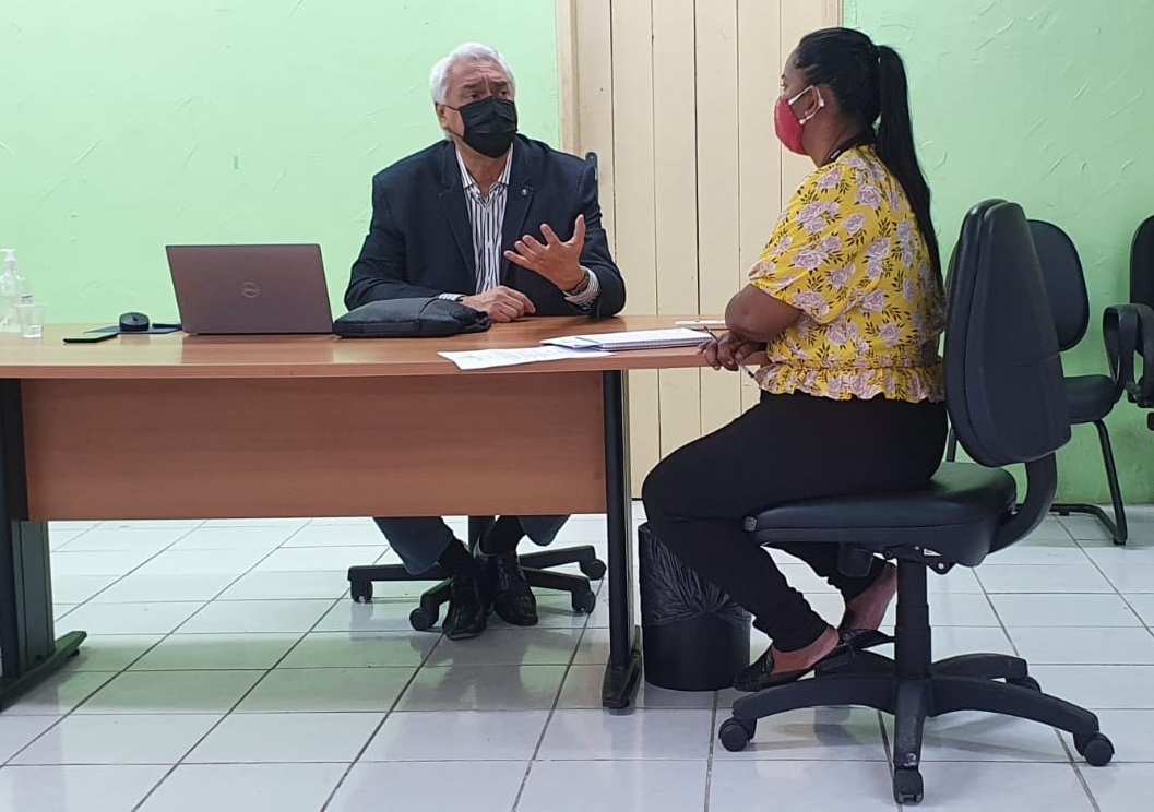 Homem de cabelo grisalho, terno preto, sentado, conversa com mulher parda, calça preta e blusa amarela, com cabelo preso.