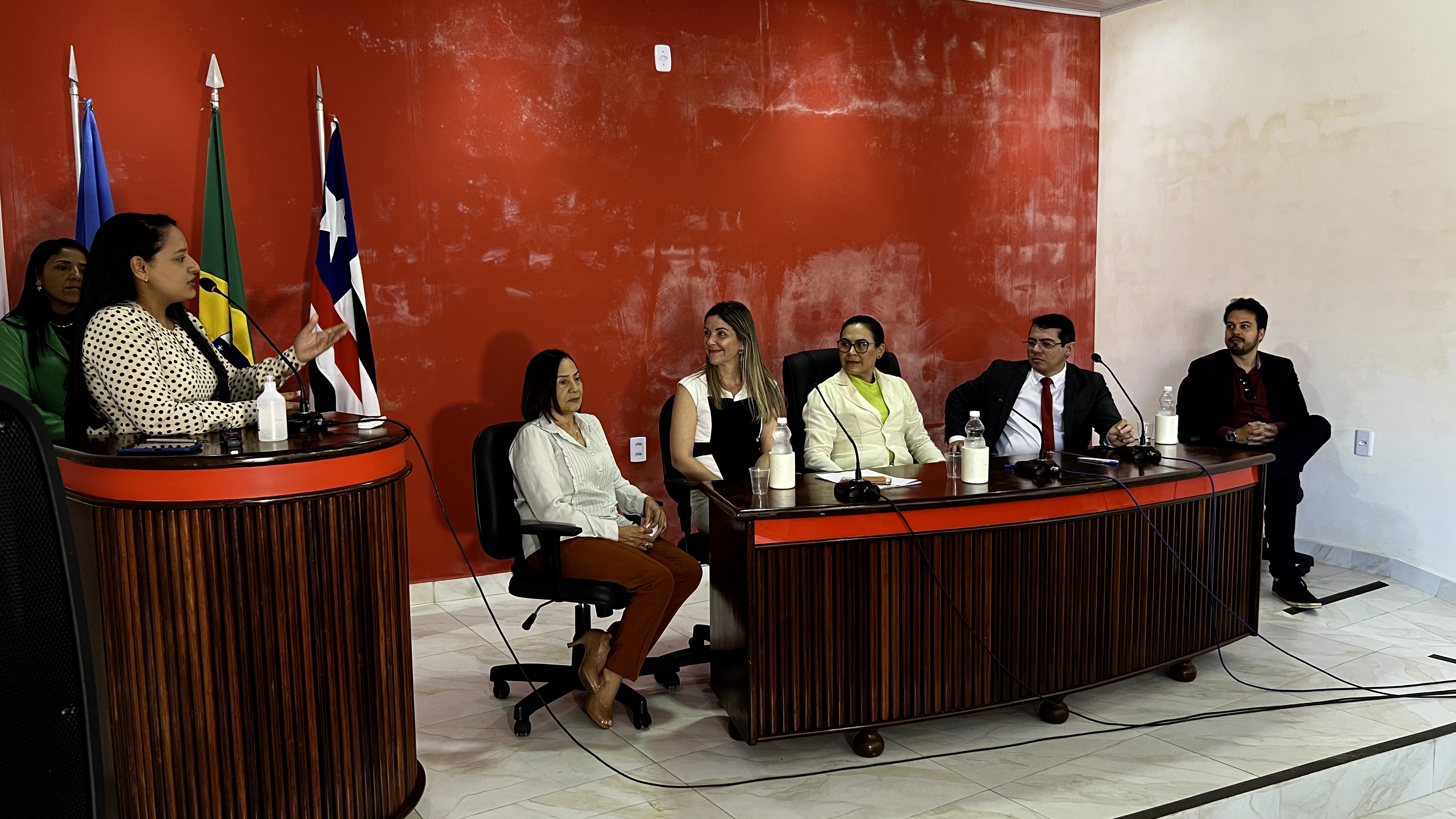 autoridades dispostas em mesa observam fala da procuradora-geral do município direto do púlpito, ao fundo parede vermelha