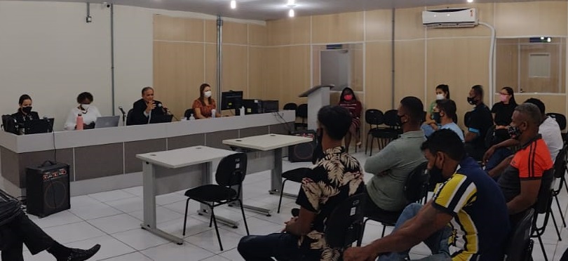 audiência na Justiça, com juiz sentado na mesa, acompanhado de funcionários e homens sentados na plateia.