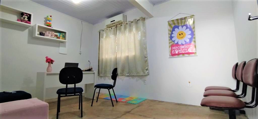 Sala de depoimento especial com decoração infantil, computador, mesa cadeira, e ar condicionado, parede branca.