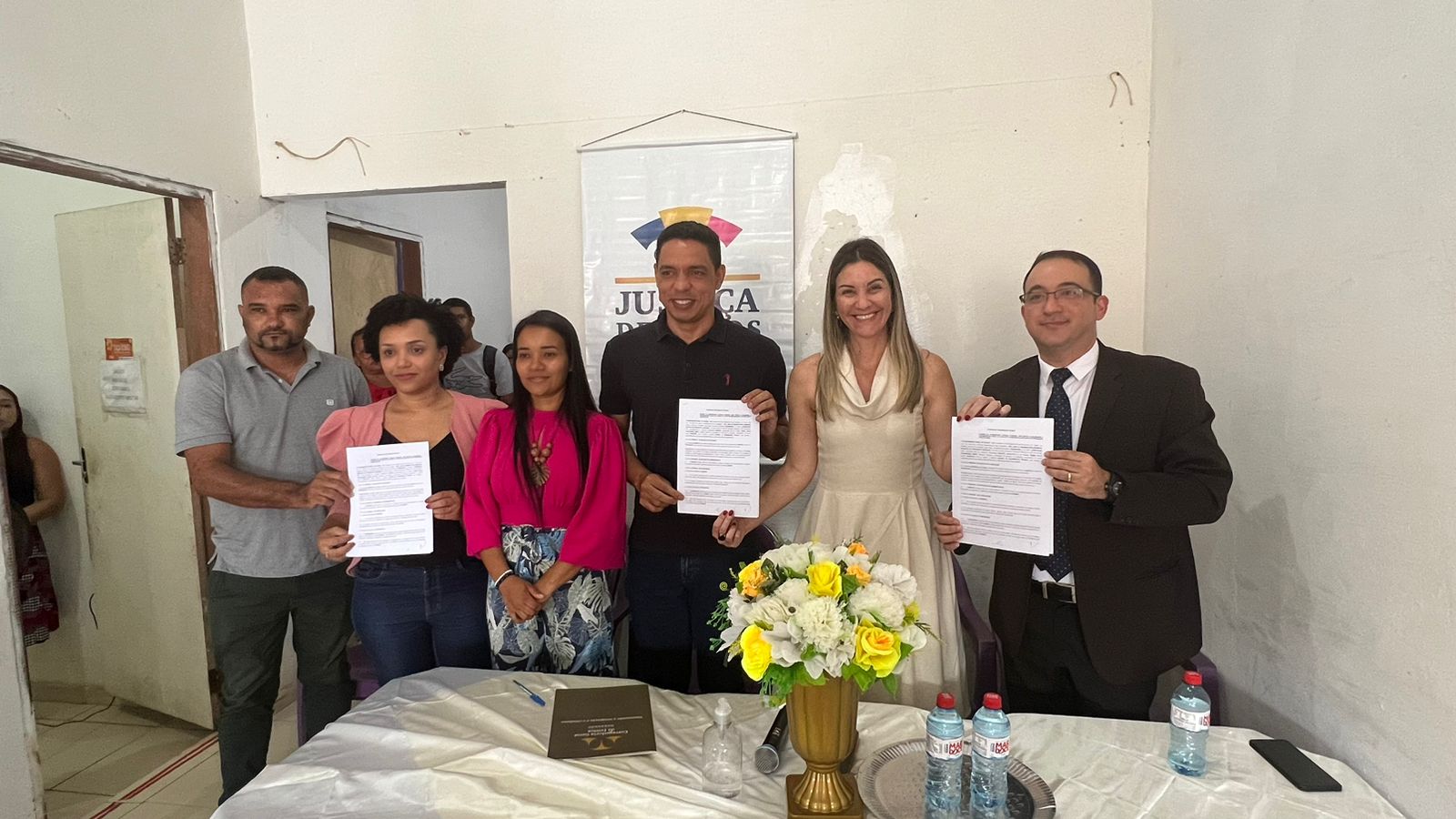 autoridades do judiciário e do município de presidente juscelino posam para foto enquanto segura termo de cooperação do justiça de todos, ao fundo parede bege e banner do projeto
