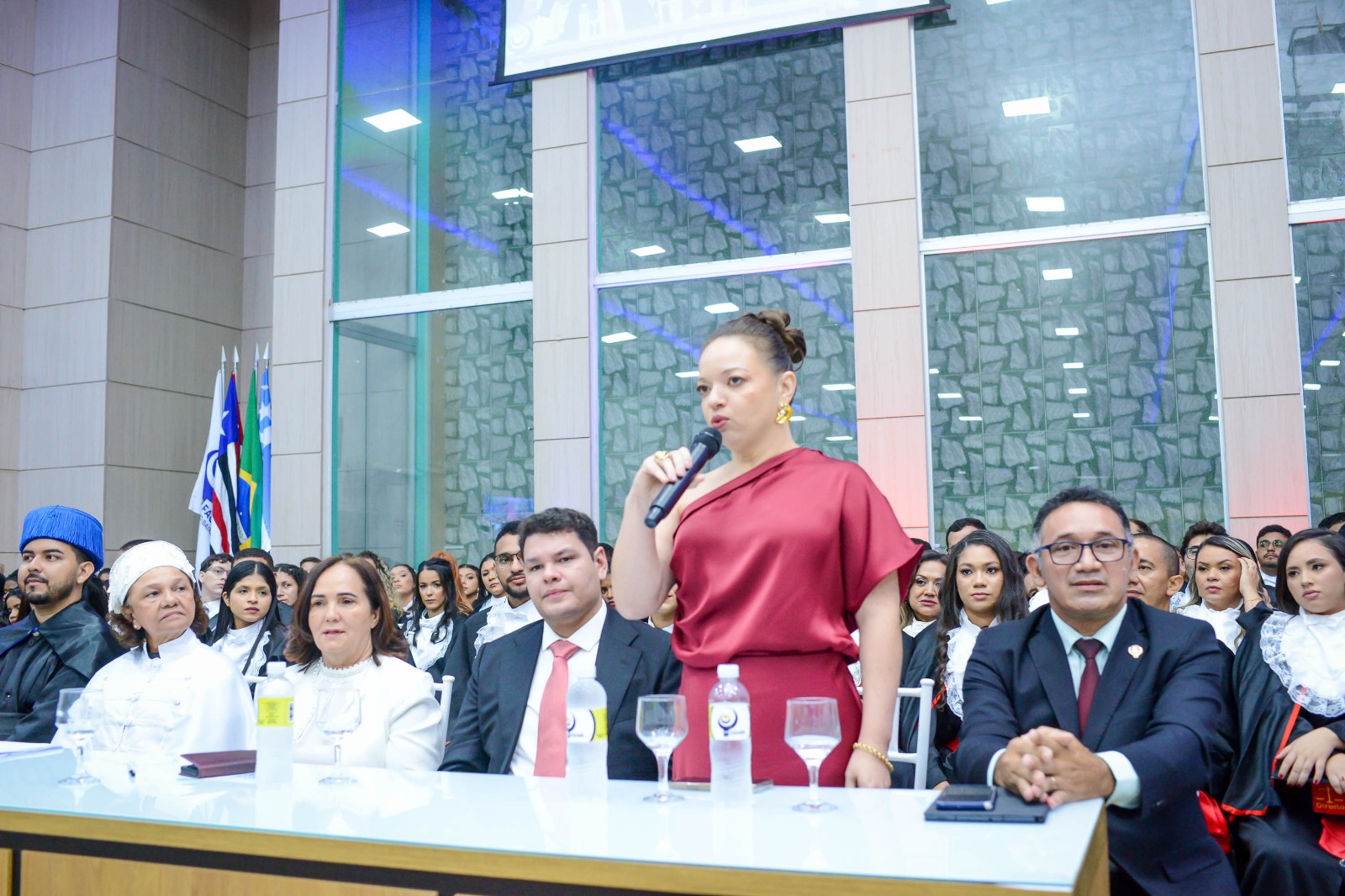 A imagem mostra uma mulher de vestido vermelho falando em um microfone em um ambiente formal. Ao seu redor, há pessoas sentadas, algumas usando trajes acadêmicos, sugerindo uma cerimônia de formatura ou acadêmica. No fundo, há bandeiras e o local parece ser um salão grande, bem iluminado, com uma parede de pedra e janelas grandes. Na mesa à frente da mulher, há vários copos e garrafas de água.