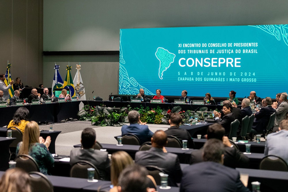 A imagem mostra uma configuração formal de reunião ou conferência, com várias pessoas sentadas ao redor de uma mesa grande disposta em formato de ¿U¿. A sala parece espaçosa, com teto alto, e há uma faixa ao fundo que diz: ¿ENCONTRO DO CONSELHO DE PRESIDENTES DOS TRIBUNAIS DE JUSTIÇA DO BRASIL - CONSEPRE - 5 A 8 DE JUNHO DE 2024 CHAPADA DOS GUIMARÃES MATO GROSSO¿. Isso indica que o evento é uma reunião do Conselho de Presidentes dos Tribunais de Justiça do Brasil, programada de 5 a 8 de junho de 2024, em Chapada dos Guimarães, Mato Grosso. A presença de bandeiras sugere representação oficial de diferentes regiões ou entidades.