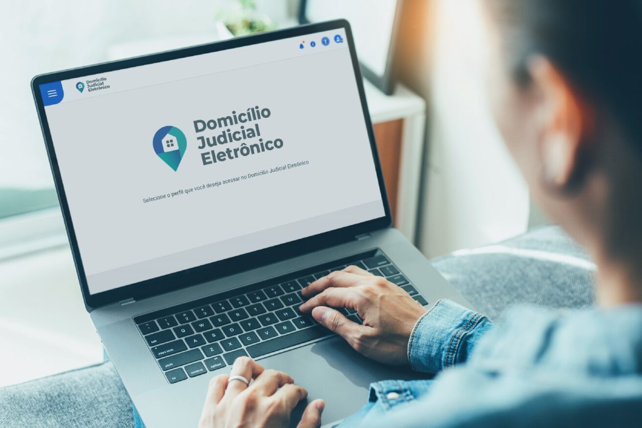 A imagem mostra uma pessoa usando um laptop com uma tela que exibe o que parece ser uma página da web para ¿Domicílio Judicial Eletrônico¿. A página apresenta um logotipo composto por uma bolha de fala azul com escalas de equilíbrio brancas dentro dela, e o texto ¿Domicílio Judicial Eletrônico¿ abaixo em azul. Também há texto adicional na tela que está muito pequeno para ler claramente. A pessoa está vestindo uma blusa cinza de mangas compridas, e apenas a parte de trás do laptop e parte do lado esquerdo da pessoa são visíveis. O cenário sugere um ambiente para trabalho jurídico ou judicial sendo conduzido online, o que pode ser relevante para quem se interessa por processos legais ou comunicação eletrônica dentro do sistema judiciário