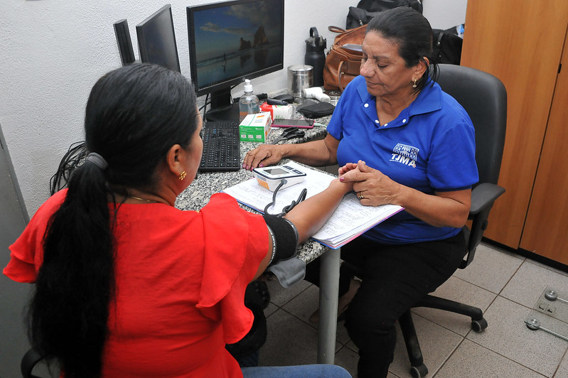 Foto em ambiente interno, com enfermeira realizando atendimento de servidora (aferição de pressão arterial)