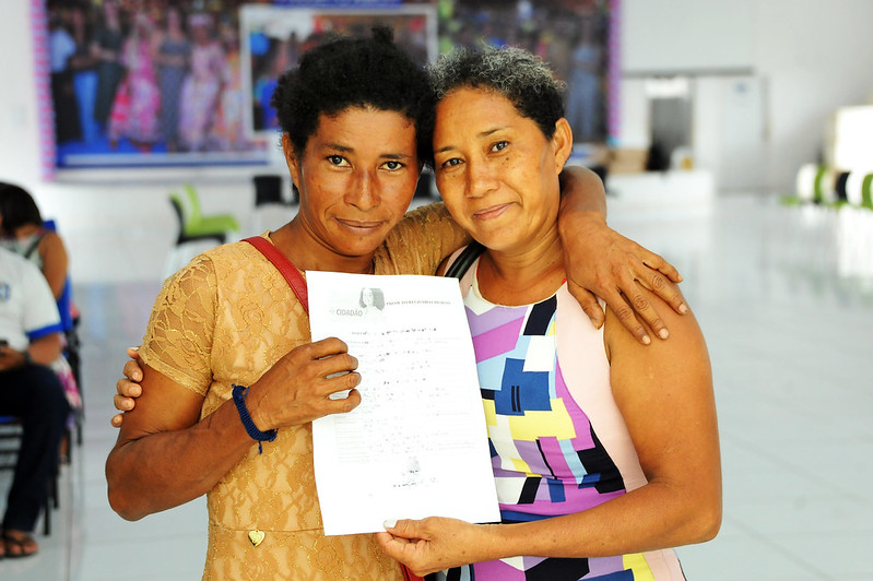 Em foto da cintura pra cima, duas mulheres abraçadas sorriem e mostram documento do projeto Registro Cidadao