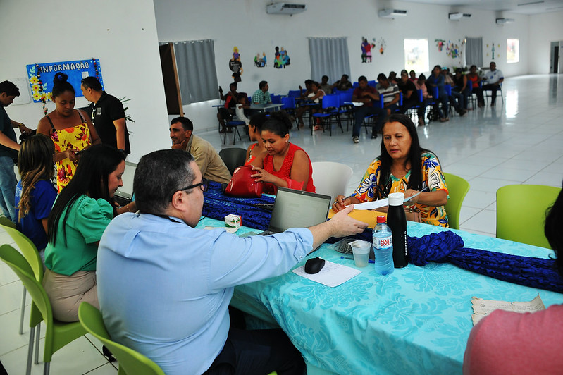 Durante projeto Registro Cidadao em Zé Doca, servidores do TJMA atendem pessoas da comunidade em mesa retangular, enquanto outras aguardam atendimento sentadas na parte de trás do salao