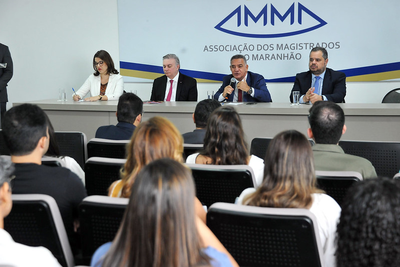 Foto em auditorio, com pessoas sentadas aparecendo de costas, e autoridades à frente na mesa do evento, com o desembargador Ronaldo Maciel fazendo uso da fala