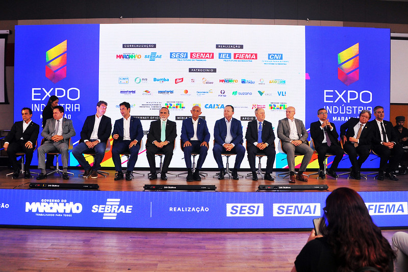 Em mesa de abertura de solenidade, com fundo de palco com logomarcas da Expo Industria, doze homens sentados lado a lado