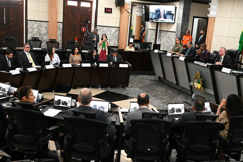 A imagem retrata uma reunião oficial ou assembleia, possivelmente em um ambiente governamental ou judicial. A sala tem um layout semicircular com um palco central elevado, onde sete indivíduos estão sentados atrás de uma longa mesa, de frente para a plateia. Cada pessoa no palco possui um microfone e papéis à sua frente. A bandeira nacional do Brasil e a bandeira da Bahia estão expostas atrás dessa mesa central, junto com uma cruz cristã acima das bandeiras.

Na frente do palco, há uma plateia composta por pessoas vestindo trajes civis e algumas em uniformes militares ou de serviços de emergência. O piso possui um padrão geométrico com azulejos contrastantes em tons escuros e claros que irradiam do centro da sala.