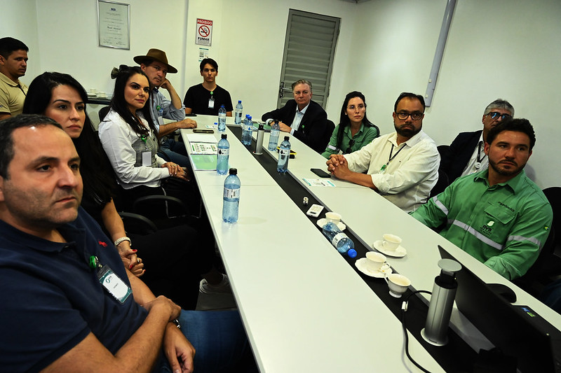 Foto em ambiente interno de sala de reunioes, com grupo de pessoas sentadas em volta de mesa retangular, com televisor mostrando imagem aérea de empresa siderúrgica. O presidente do TJMA, desembargador Froz Sobrinho, está na ponta