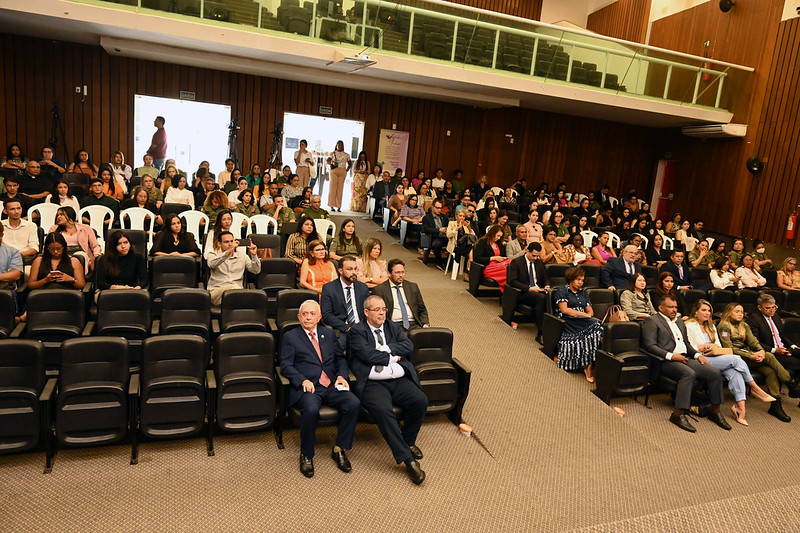 Foto de auditório com carpete cinza e poltronas pretas. A maior parte dos lugares está ocupada com pessoas vestidas formalmente e algumas com farda de forças de segurança