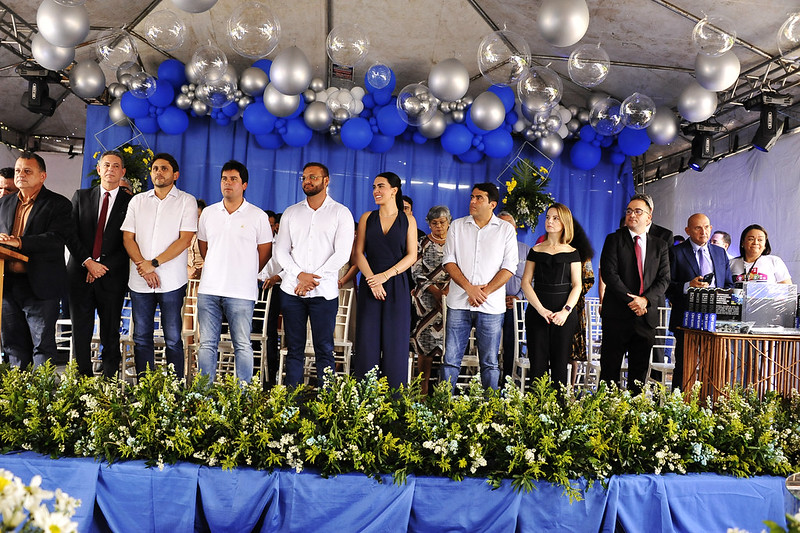 A imagem mostra um grupo de pessoas reunidas em um palco decorado com balões azuis e prateados, além de arranjos florais. O fundo do palco é azul e há uma cobertura acima dele. Os participantes estão vestidos com roupas formais ou semi-formais, indicando que se trata de um evento ou cerimônia formal.

Na frente do palco, há arranjos florais verdes e brancos, e cadeiras estão dispostas atrás das pessoas. Também é visível um púlpito no lado esquerdo, sugerindo que o evento pode incluir discursos ou apresentações. É um belo registro de uma celebração ou encontro oficial, com atenção aos detalhes na decoração e disposição dos participantes.
