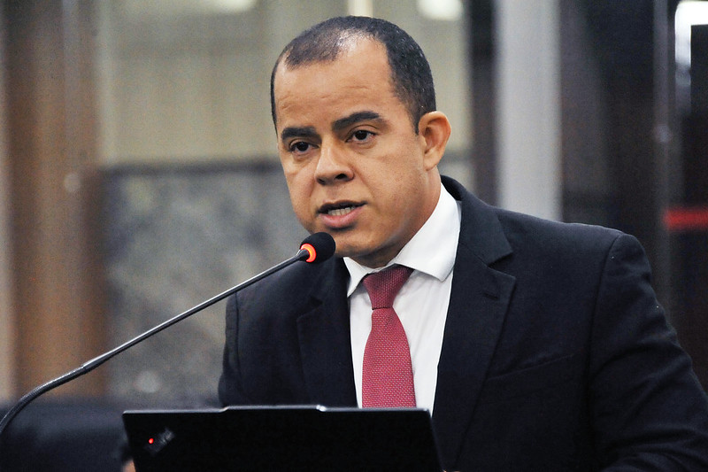 homem vestindo terno escuro falando ao microfone, durante reuniao