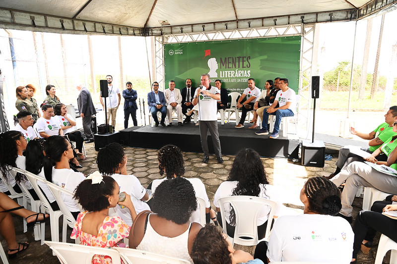 A imagem mostra um grupo de pessoas reunidas sob uma tenda para um evento. No palco, há várias pessoas sentadas, enquanto uma está de pé e fala ao microfone. A plateia, composta por participantes sentados em cadeiras plásticas brancas, acompanha atentamente a apresentação.O fundo do palco possui um banner verde com os dizeres "MENTES LITERÁRIAS" e "Projeto Jovens Arte e Design", indicando que o evento está relacionado à literatura, arte e design, possivelmente envolvendo jovens.