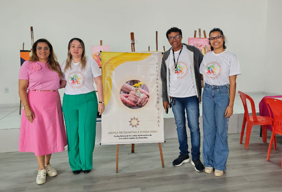Na imagem, quatro pessoas posam sorrindo em um ambiente interno. Três delas vestem camisetas brancas com o logotipo do Nejur e uma veste um vestido rosa. Ao centro, há um banner com o título ¿Justiça Restaurativa e Pessoa Idosa¿ e o brasão do Tribunal de Justiça do Maranhão. Ao fundo, vê-se parte da decoração com quadros coloridos em cavaletes.