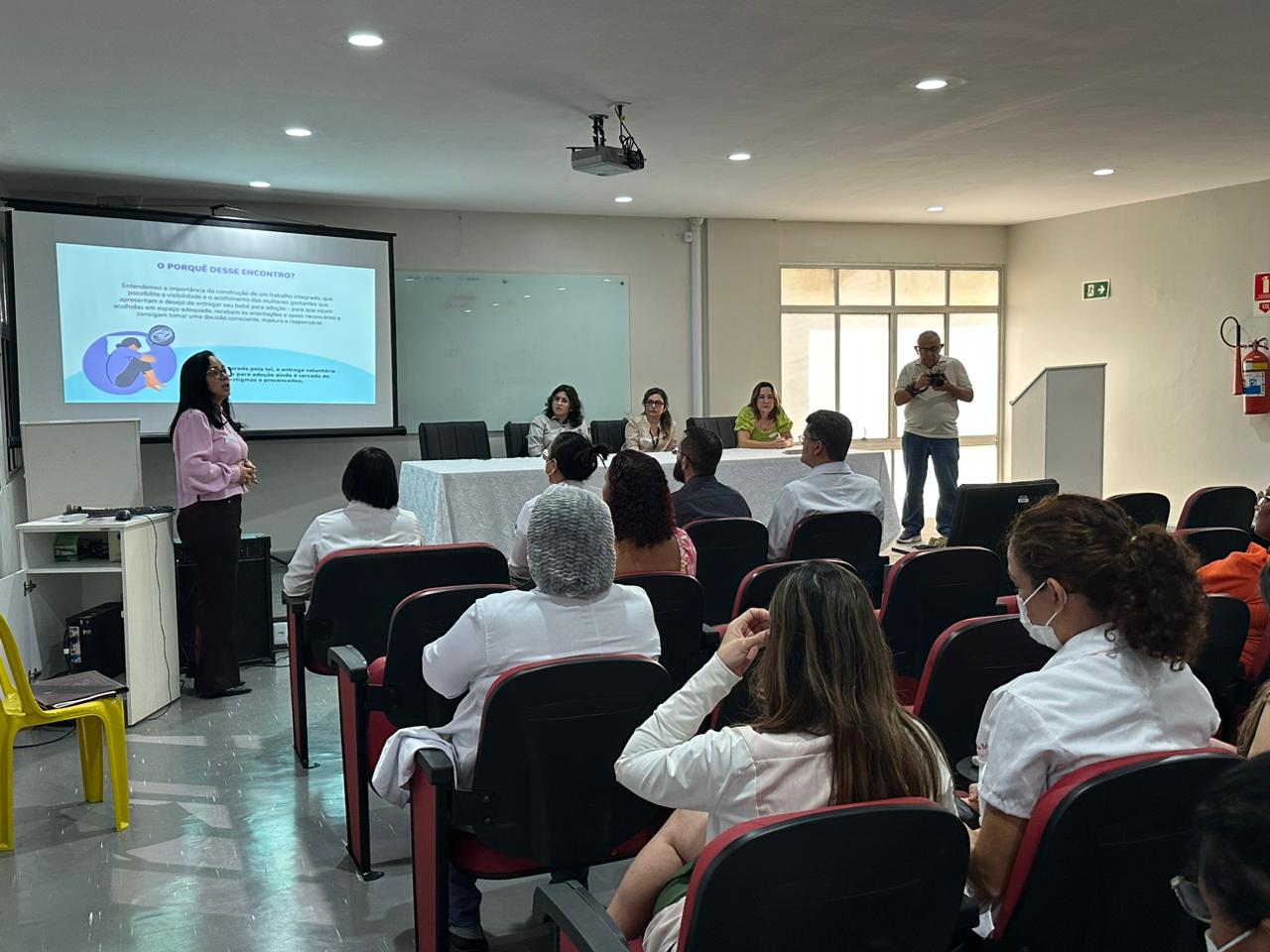 Foto de uma sala de reuniões com um telão passando slides uma senhora em pé fazendo a apresentação, duas senhoras sentadas na mesa de autoridades e pessoas sentadas assistindo a palestra