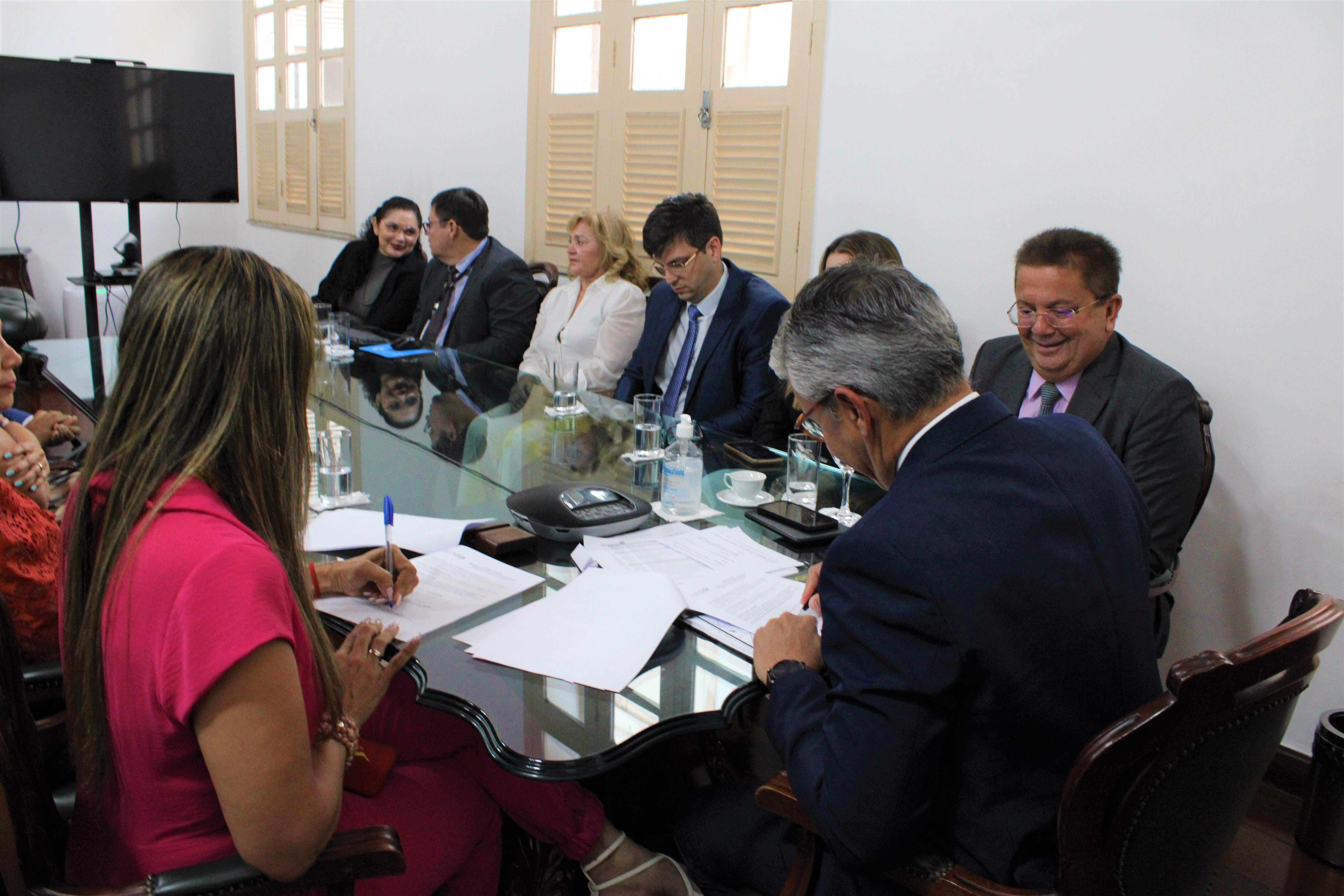 Pessoas sentadas ao redor de mesa, na cabeceira, o presidente do TJMA, desembargador Paulo Velten assina documento e a diretora-presidente do CRF-MA assina na esquerda.