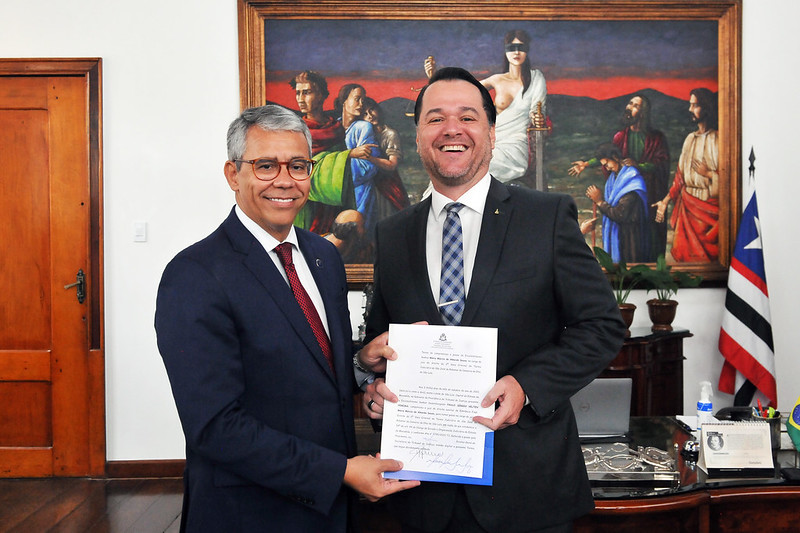 Foto do presidente do TJMA, desembargador Paulo Velten de terno azul escuro e gravata vinho ao lado do juiz promovido, alto, branco, de terno escuro e gravata azul e branca, sorridentes com a pasta na mão.