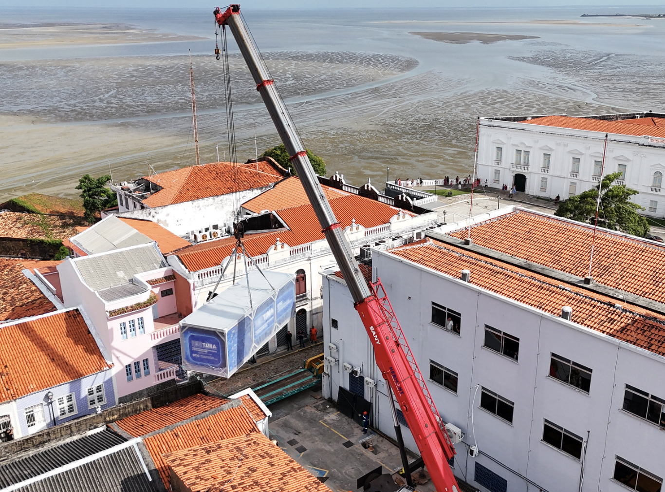 Fotografia aérea mostrando a instalação do novo Data Center no complexo do Edifício Durval Soares da Fonseca, em uma área histórica de São Luís. Um grande guindaste vermelho ergue o contêiner, que está suspenso por cabos sobre uma área interna do prédio. Ao redor, aparecem edifícios históricos com fachadas brancas e telhados de cerâmica alaranjada. Ao fundo, é possível ver a paisagem da região costeira, com o mar e áreas de mangue durante a maré baixa.