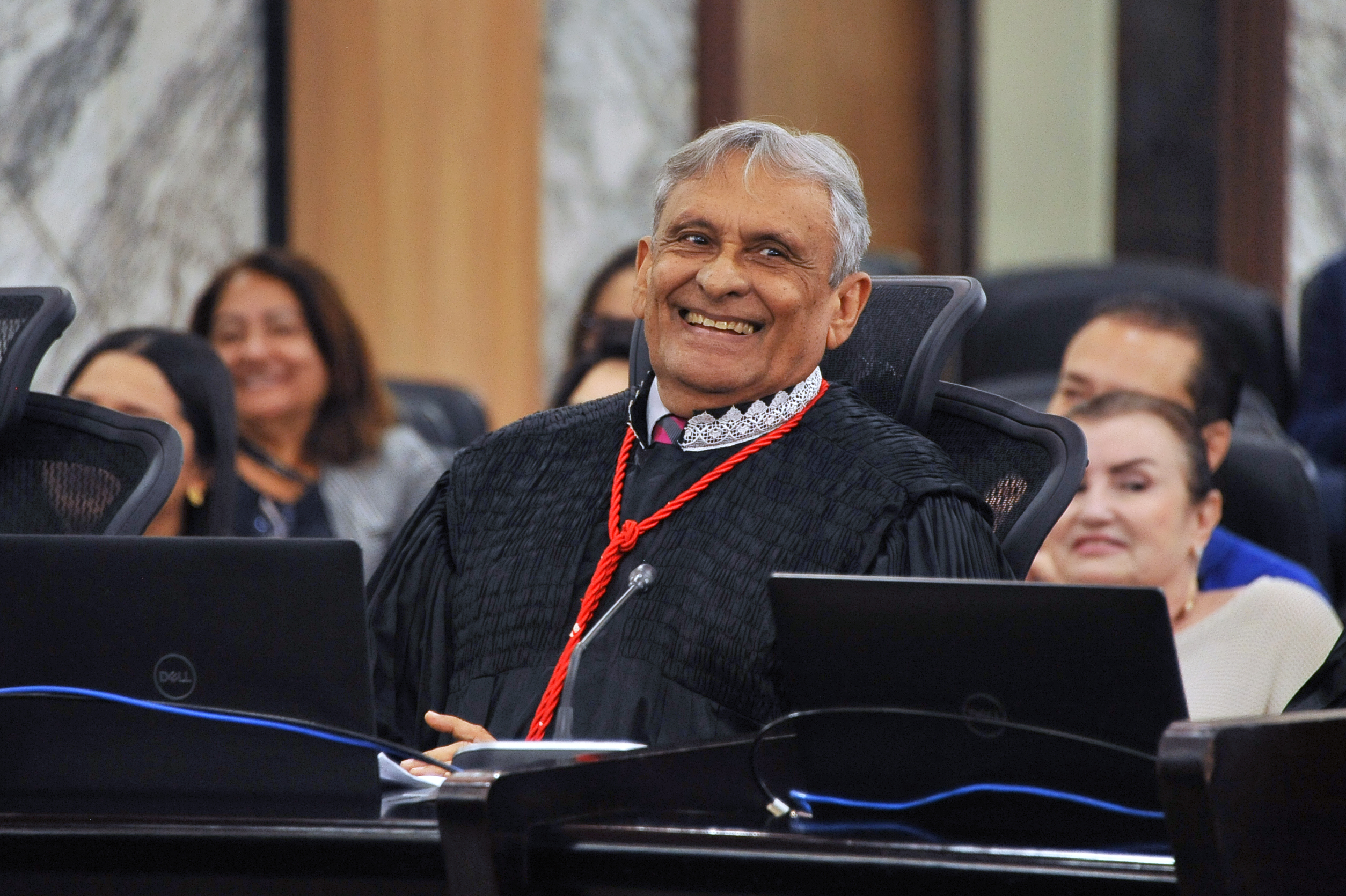 Homem de pele escura, cabelos grilhasos e idade senil, usando toga e sorrindo, sentado na cadeira de desembargadores do Pleno do Tribunal de Justiça. Ao fundo, do lado direito, mulher sentada rindo.