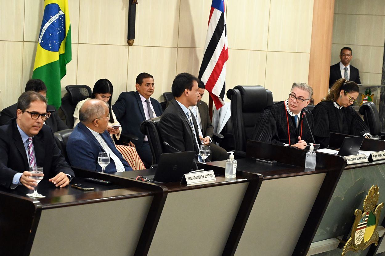 A imagem mostra uma reunião formal em uma sala de conferências. Há várias pessoas sentadas em uma mesa longa, algumas delas usando trajes formais, incluindo togas, sugerindo que pode ser uma sessão de um tribunal ou uma reunião de um conselho jurídico. Na mesa, há placas de identificação, copos de água, laptops e um frasco de álcool em gel. Ao fundo, há duas bandeiras: a bandeira do Brasil e a bandeira do estado do Maranhão. A parede ao fundo é revestida de madeira clara, e há um crucifixo pendurado na parede. A placa de identificação mais visível na mesa diz "PROCURADOR DE JUSTIÇA".