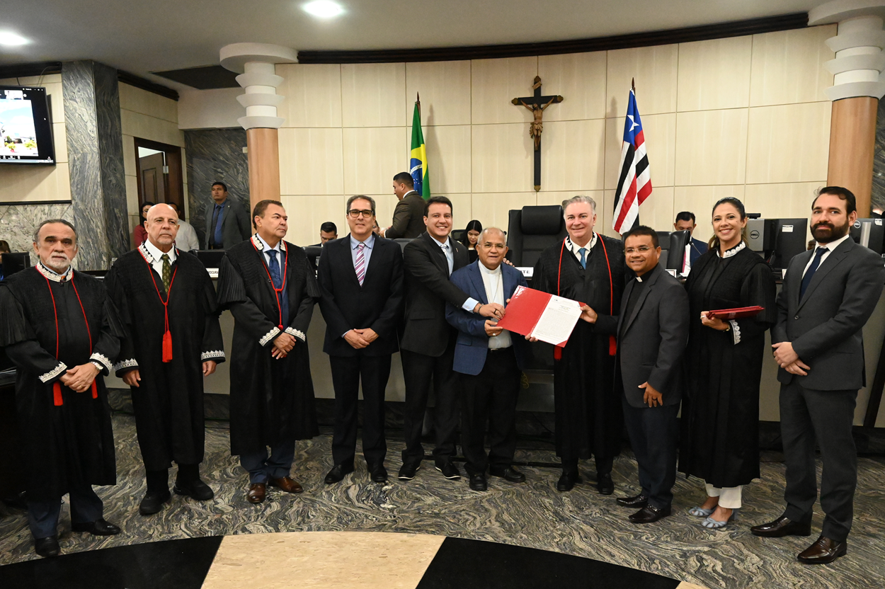 A imagem mostra um grupo de pessoas em um ambiente formal, possivelmente um tribunal ou sala de reunião oficial. Os indivíduos estão vestidos com trajes formais, alguns usando togas judiciais e outros ternos. Estão em frente a uma parede com um crucifixo e duas bandeiras, uma do Brasil e a outra do estado do Maranhão. O grupo segura uma pasta ou documento vermelho, que parece ser o foco da reunião. O cenário sugere um evento oficial ou cerimonial, possivelmente relacionado ao judiciário ou ao governo.