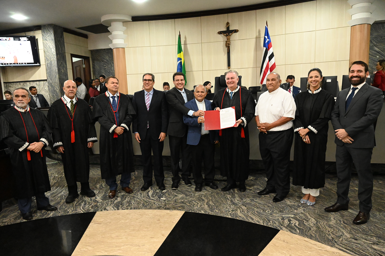 A imagem mostra um grupo de pessoas em um ambiente formal, possivelmente um tribunal ou sala de reunião oficial. Os indivíduos estão vestidos com trajes formais, alguns usando togas judiciais e outros ternos. Estão em frente a uma parede com um crucifixo e duas bandeiras, uma do Brasil e a outra do estado do Maranhão. O grupo segura uma pasta ou documento vermelho, que parece ser o foco da reunião. O cenário sugere um evento oficial ou cerimonial, possivelmente relacionado ao judiciário ou ao governo.