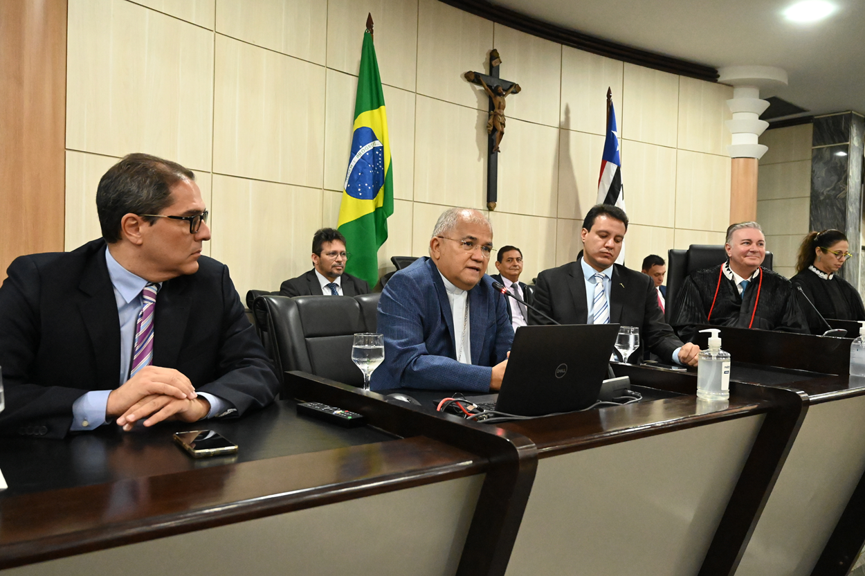 A imagem mostra uma reunião formal em uma sala de conferências. Há várias pessoas sentadas em uma mesa, algumas usando ternos e outras vestindo togas, sugerindo que pode ser uma reunião judicial ou governamental. Na mesa, há um laptop, copos de água, um frasco de álcool em gel e alguns controles remotos. Ao fundo, há duas bandeiras: uma do Brasil e outra de um estado brasileiro. Também há um crucifixo na parede.