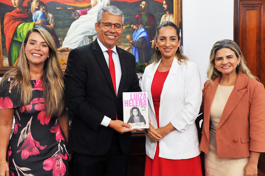 Foto posada com as três visitantes e o desembargador paulo velten em pé, lado a lado. o desemnargador mostra a capa do livro "Luiza Helena:mulher do Brasil"