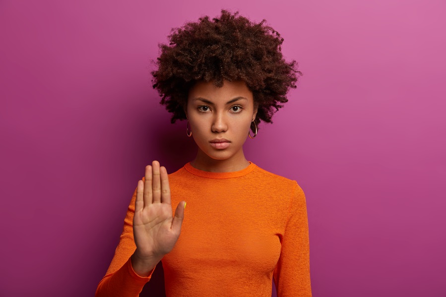 Mulher negra, com cabelos médios e encaracolados, olha séria para frente e levanta a mão direita em sinal de pare. Ela usa camisa de manga comprida laranja. Fundo da foto é rosa escuro.