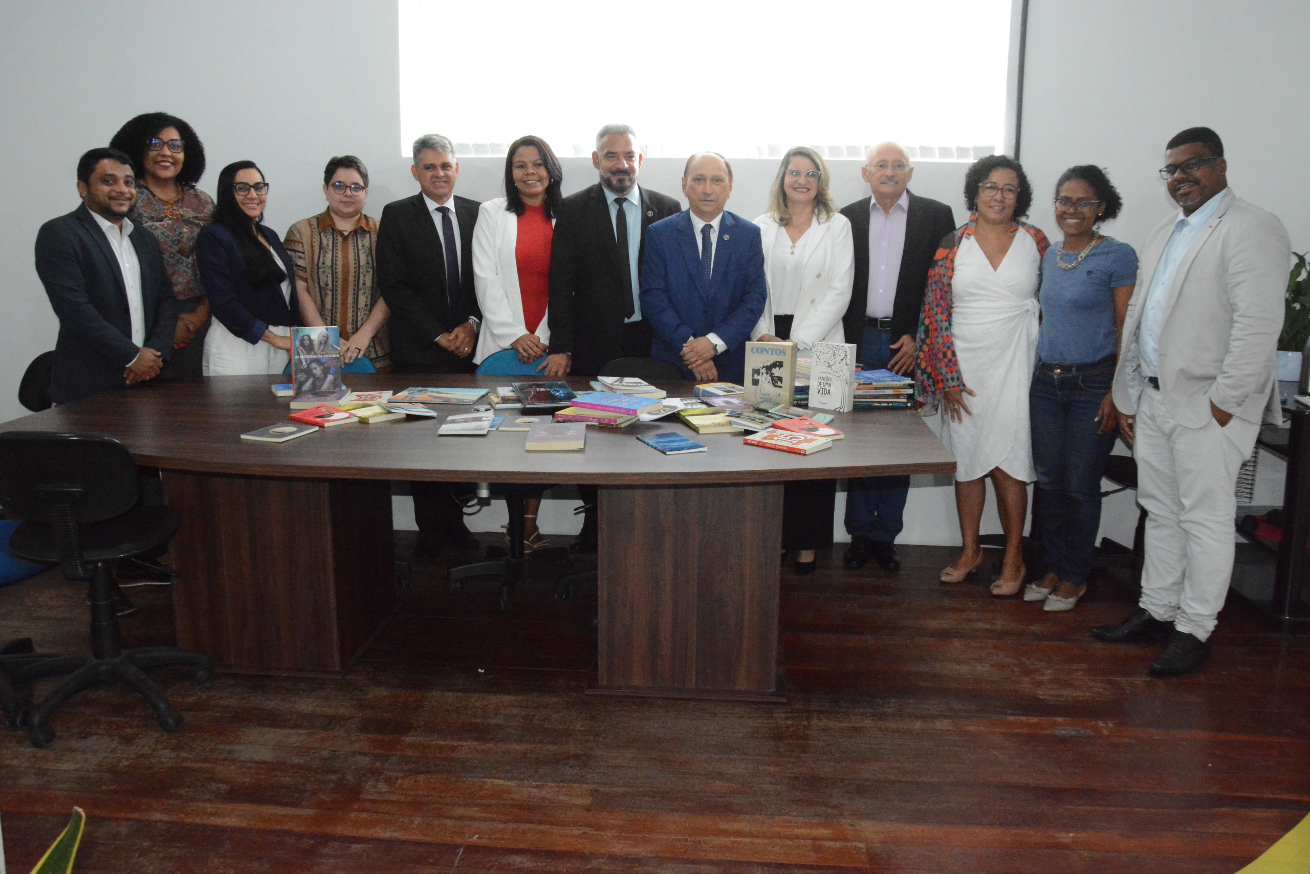 grupo de pessoas em pé lado a lado, vestidos de forma formal e casual, sendo homens e mulhers, em ambiente interno atrás de uma mesa