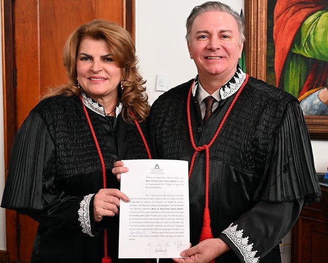 A imagem mostra dois magistrados de pé, lado a lado, vestindo togas judiciais pretas com detalhes em vermelho e branco e segurando o termo de posse. Ao fundo, há uma parede de madeira e uma pintura colorida pendurada