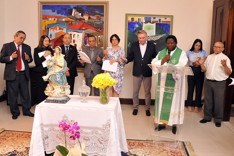 Foto de magistrados e magistradas juntos em pé atrás de uma mesa com a imagem de Nossa Senhora da Conceição em momento religioso com o padre falando