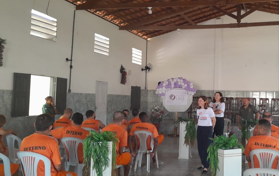 Foto em ambiente interno de sala de aula, com internos vestindo camisa do sistema prisional de costas, e à frente deles servidora da CEMULHER falando em pé