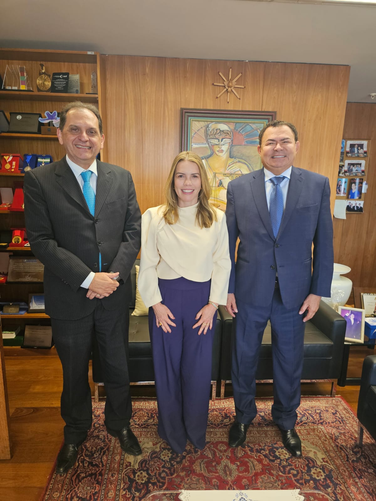 Foto vertical, colorida, de uma mulher entre dois homens, de pé, em ambiente interno com móveis de madeira e uma pintura na parede ao fundo. As duas pessoas nas extremidades estão vestindo ternos escuros, enquanto a pessoa no centro está vestindo uma blusa branca e calças azuis. A sala tem teto branco e tapete vermelho estampado.Ao fundo, sofá escuro.