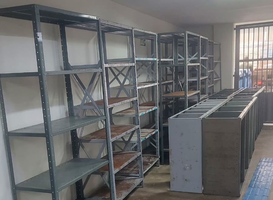 Foto horizontal, colorida, mostra fileiras de estantes de aço em sala, junto a uma prede branca. Ao fundo, uma porta fechada com grade.