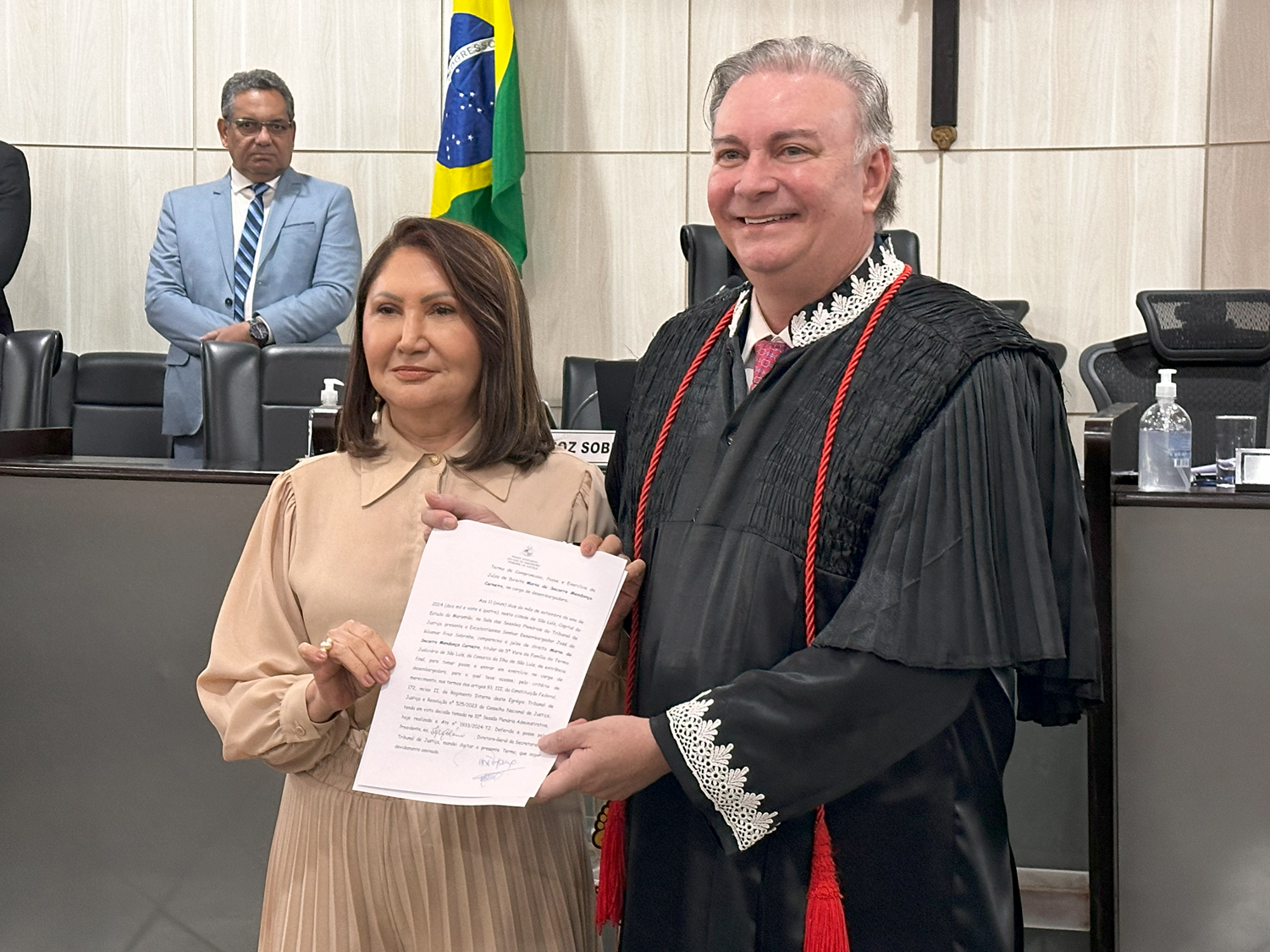 A imagem mostra três pessoas em um ambiente formal de salão de um tribunal. No centro, a desembargadora Maria do Socorro durante solenidade de posse, ao lado do desembargador Froz Sobrinho, usando vestes judiciais com rendas nas mangas e segurando um documento. Eles estão em frente a uma mesa com microfones e uma bandeira do Brasil ao fundo. O documento segurado pela pessoa com vestes judiciais sugere que algum tipo de procedimento legal ou oficial está acontecendo.