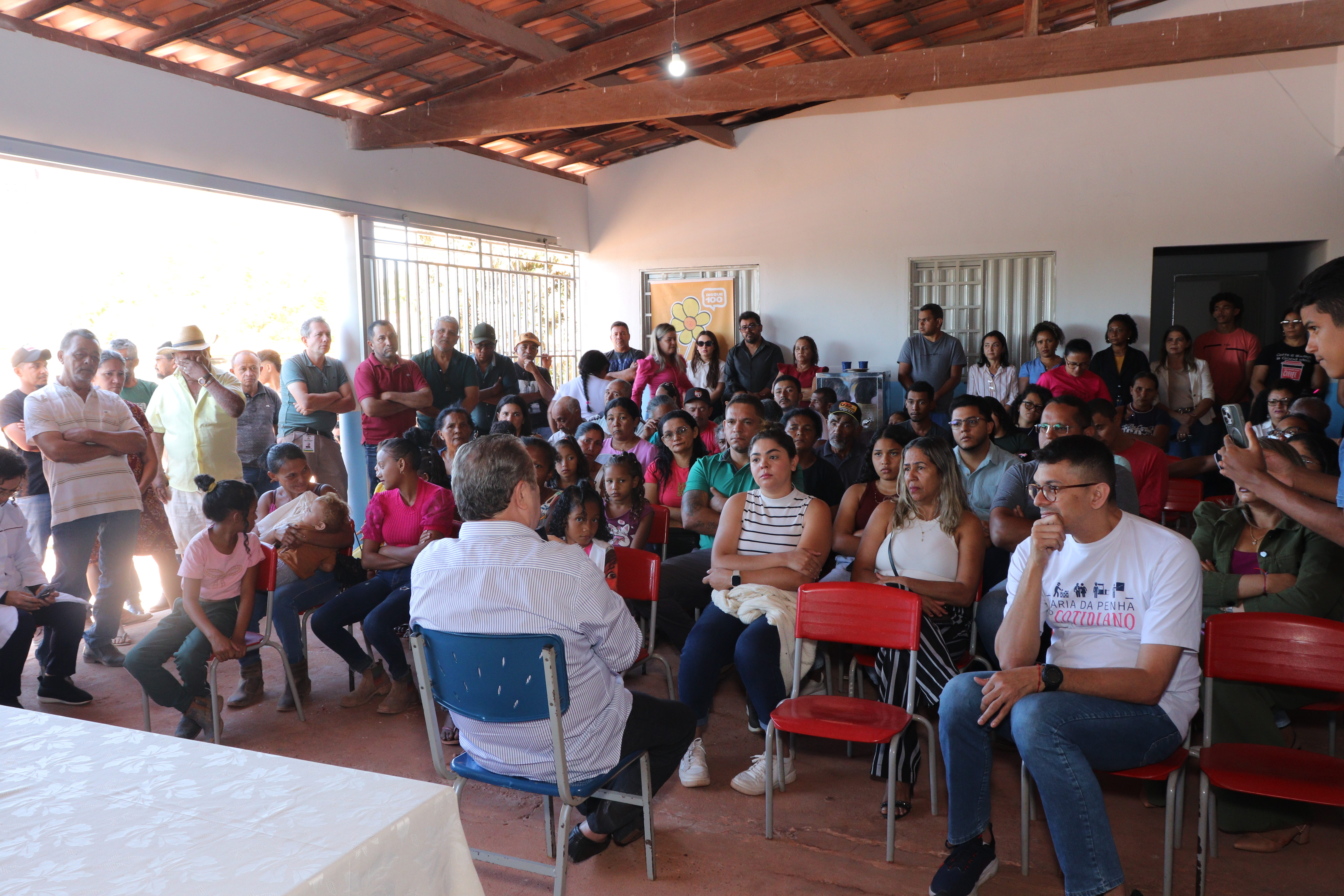 A imagem mostra uma **reunião** ou **evento comunitário** em um local coberto, com telhas de barro no teto e vigas de madeira aparentes. O ambiente está cheio de pessoas sentadas em cadeiras de plástico vermelhas, assistindo a uma apresentação ou discussão.
No centro da frente da foto, um homem de costas para a câmera, com camisa listrada, está sentado em uma cadeira azul, conversando com outro homem. Este último, de frente para a câmera, veste uma camisa branca com a estampa "**DA PENHA O COTIDIANO**", a mesma que aparece em um banner na imagem anterior. Ele gesticula enquanto fala, e há um microfone na sua frente.
A **plateia**, composta por adultos e algumas crianças, presta atenção. Pessoas em pé também cercam a área da reunião. O cenário indica um evento de caráter social ou educacional, possivelmente relacionado a um projeto ou iniciativa.