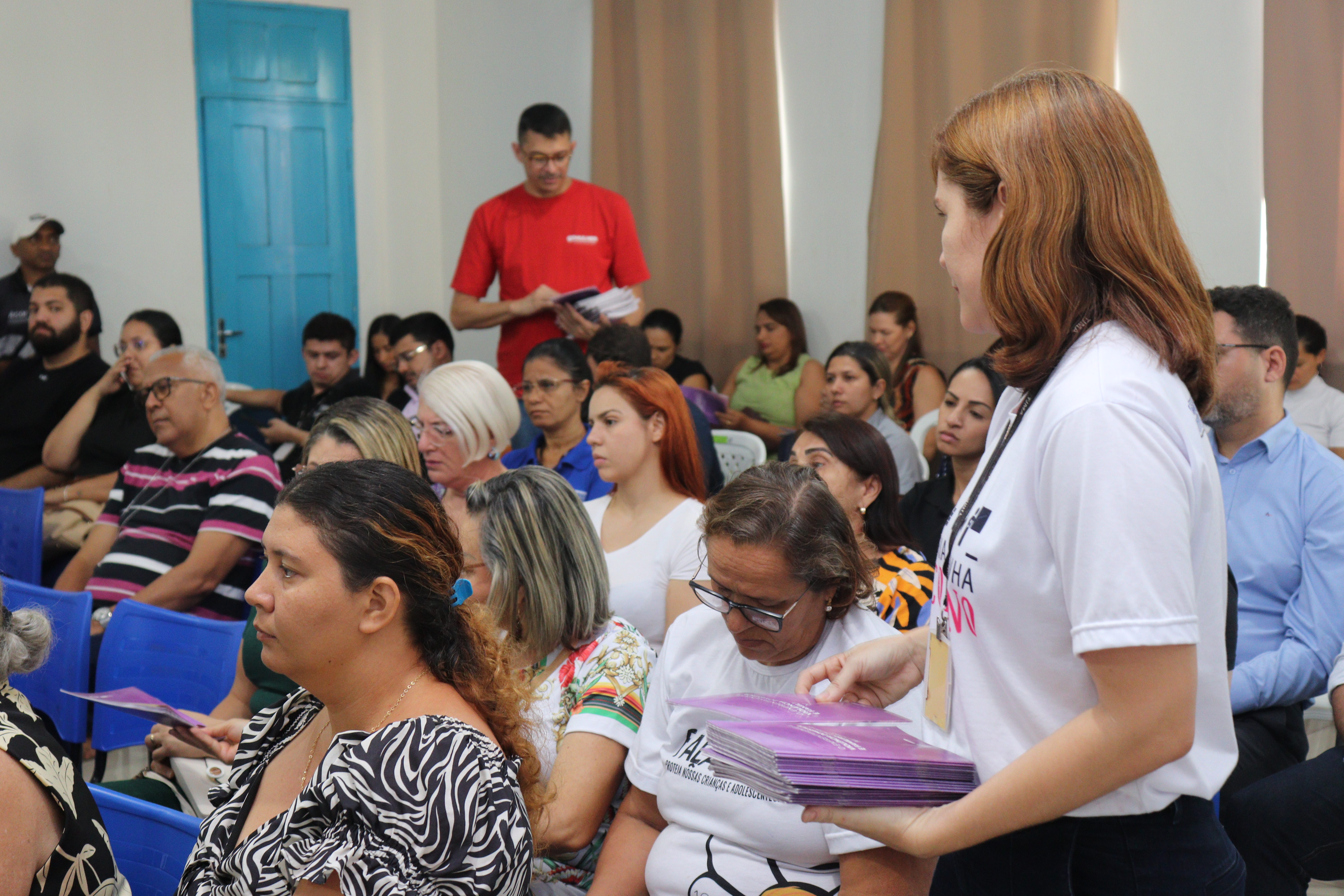 A imagem mostra um evento ou palestra em andamento. Em primeiro plano, no lado direito, uma mulher de cabelo ruivo/castanho claro, vestindo uma camiseta branca com uma estampa sutil no peito, está de pé e distribuindo folders roxos para o público. Ela está de perfil para a câmera, com o olhar voltado para as pessoas na primeira fileira.
A **plateia** é composta por dezenas de pessoas de todas as idades, a maioria sentada em cadeiras azuis. Um homem de camiseta vermelha está no fundo, em pé, também parecendo estar distribuindo materiais.
A cena sugere que o evento está em pleno andamento, com a organização distribuindo informações para os participantes. O ambiente tem uma porta azul no fundo e cortinas laranjas, semelhantes às vistas em outra imagem.