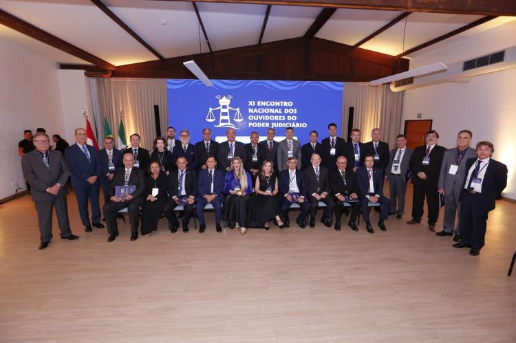 A imagem mostra um grupo de pessoas posando para uma foto em um evento formal. O fundo da imagem tem um banner azul com o texto "XI ENCONTRO NACIONAL DOS OUVIDORES DO PODER JUDICIÁRIO" e um símbolo de balança de justiça. As pessoas estão vestidas formalmente, a maioria usando ternos e gravatas.  Há bandeiras ao lado esquerdo da imagem.