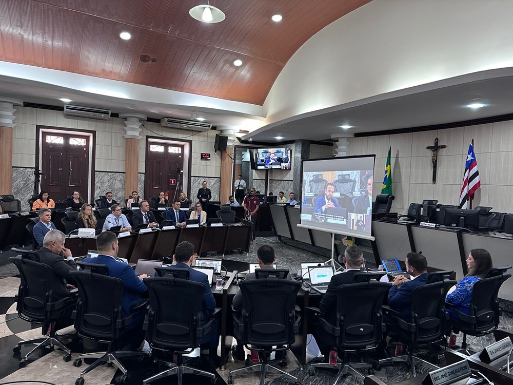 A imagem mostra a sala de sessoes do TJMA com várias pessoas sentadas em formato de ¿U¿. A sala tem um design clássico, com painéis de madeira e colunas de mármore. No centro, há uma grande tela exibindo o que parece ser a mesma reunião, sugerindo uma configuração de videoconferência. Os participantes estão vestidos com trajes profissionais, indicando um ambiente corporativo ou governamental. Documentos e laptops estão sobre a mesa, o que sugere que esta pode ser uma assembleia de negócios.