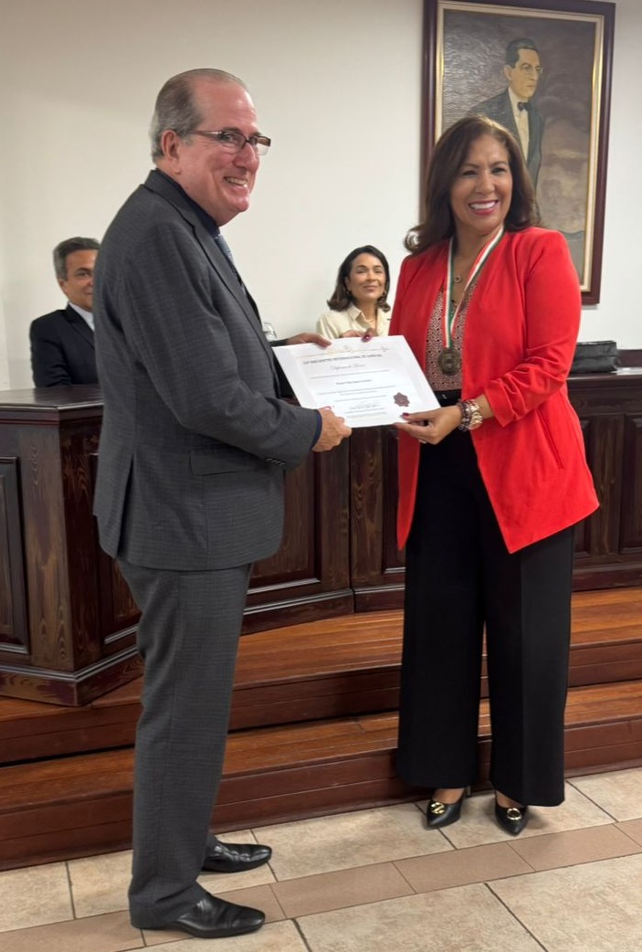 A imagem mostra uma pessoa em um terno cinza entregando um certificado para outra pessoa que está vestindo uma jaqueta vermelha e calças pretas. A pessoa que está recebendo o certificado também está usando uma medalha ao redor do pescoço. Ao fundo, há outras três pessoas sentadas em uma mesa de madeira, e um retrato emoldurado está pendurado na parede. O cenário parece ser um evento formal, possivelmente uma cerimônia de premiação. A imagem capta um momento de reconhecimento ou conquista.
