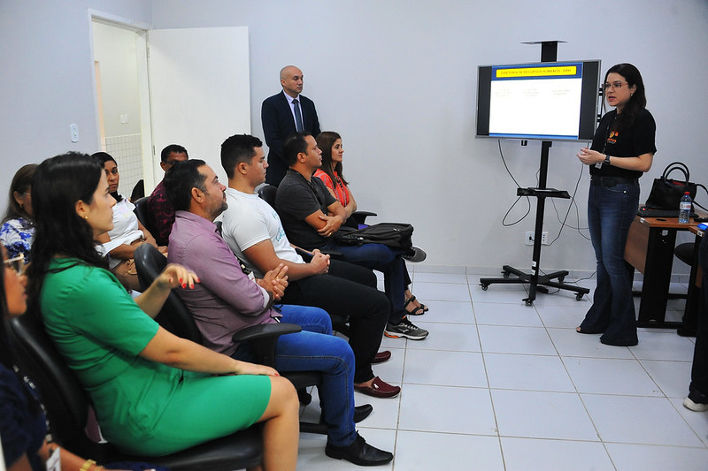 Foto horizontal do Programa Justiça de Proximidade em Paulo Ramos, palestra RH Itinerante. Em pé mulher branca, de cabelos castanhos, usa óculos de grau, blusa preta e calça jeans. Ao lado dela, um monitor de TV. De perfil na foto, sentados e sentadas, homens e mulheres de idades variadas e roupas de cores diversas. O piso e as paredes da sala são brancas. Ao fundo, há um homem em pé, calvo, branco, que usa terno.