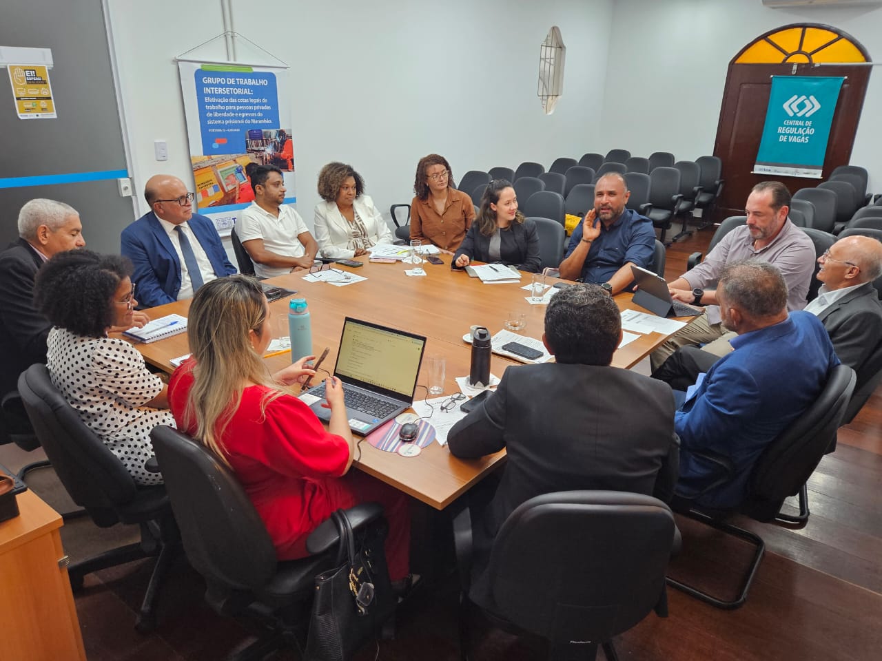 A imagem mostra uma reunião de trabalho com várias pessoas sentadas ao redor de uma mesa de conferência. Na mesa, há laptops, garrafas de água, papéis e canetas. As pessoas estão envolvidas em uma discussão, possivelmente sobre um tema profissional ou institucional. Na parede ao fundo, há dois cartazes: um à esquerda com o título "GRUPO DE TRABALHO INTERSETORIAL" e outro à direita com o título "CENTRAL DE REGULAÇÃO DE VAGAS". A reunião parece estar ocorrendo em um ambiente formal, possivelmente em um escritório ou sala de conferências.