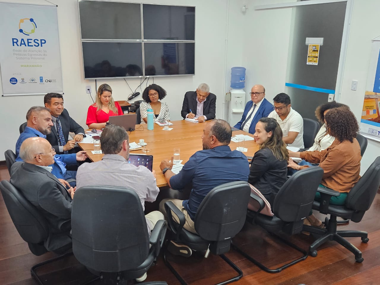 A imagem mostra uma reunião de trabalho com várias pessoas sentadas ao redor de uma mesa de conferência. Na mesa, há laptops, garrafas de água, papéis e canetas. As pessoas estão envolvidas em uma discussão, possivelmente sobre um tema profissional ou institucional. Na parede ao fundo, há dois cartazes: um à esquerda com o título "GRUPO DE TRABALHO INTERSETORIAL" e outro à direita com o título "CENTRAL DE REGULAÇÃO DE VAGAS". A reunião parece estar ocorrendo em um ambiente formal, possivelmente em um escritório ou sala de conferências.